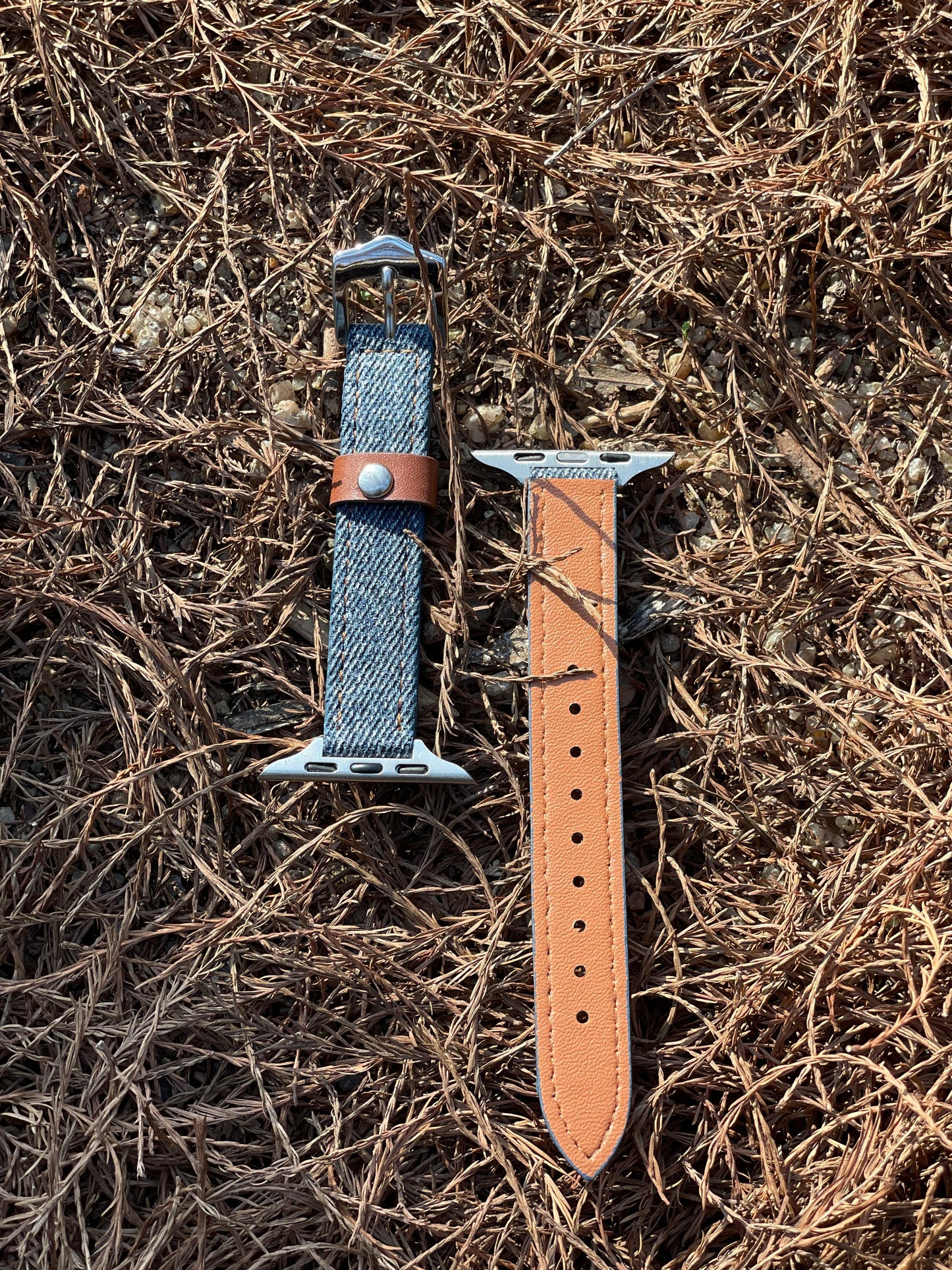 Two smartwatch straps, one side is made of leather, and the other side is made of grey-blue denim with a brown accent, on the grass. 